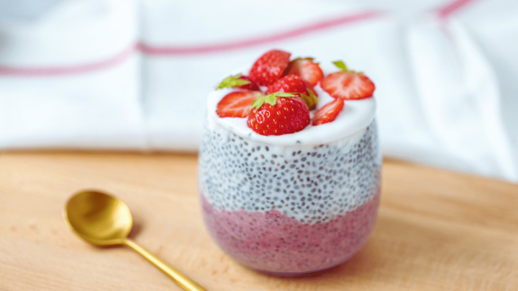 A glass filled with layered chia pudding, the bottom half showcasing a rich, creamy texture indicative of well-soaked chia seeds. The top layer is adorned with vibrant, freshly sliced strawberries, their juicy redness contrasting beautifully against the neutral tone of the pudding. The glass's transparency allows one to observe the distinct layers, making this not just a nutritious treat, but also a visual delight.
