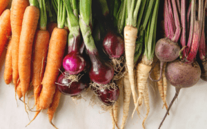 A colorful assortment of root vegetables arranged on a wooden cutting board. The vegetables include orange carrots, creamy-colored parsnips, beige turnips, and purple beets. They are assorted in a visually appealing manner, with their natural shapes and sizes on display.