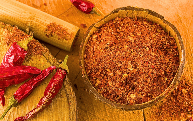 A bowl containing both whole and ground cayenne pepper, accompanied by a wooden masher, all positioned on a rustic wooden table.