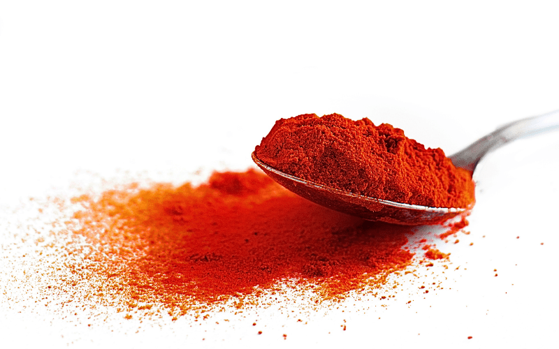 A close-up image showcases a generous portion of vibrant paprika powder delicately arranged on a spoon, with a spillage of the spice accentuating the composition against a pristine white background. The rich, earthy tones of the paprika emanate warmth and depth, while the granular texture is discernible in the finely ground powder. The intricate details of the spice, as well as the contrast between the spoon and the spilled paprika, underscore its sensory appeal and culinary significance.