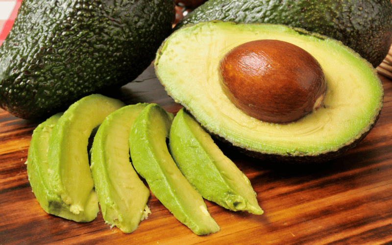 A selection of ripe avocados arranged on a natural wooden table, including whole avocados, halved avocados with the pit, and expertly sliced avocado segments. 