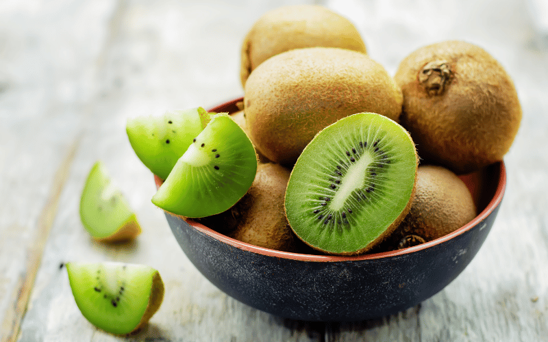 A black bowl containing whole and halved fresh kiwis, neatly arranged on a wooden table surface, with ample natural lighting emphasizing the vibrant colors of the fruit.