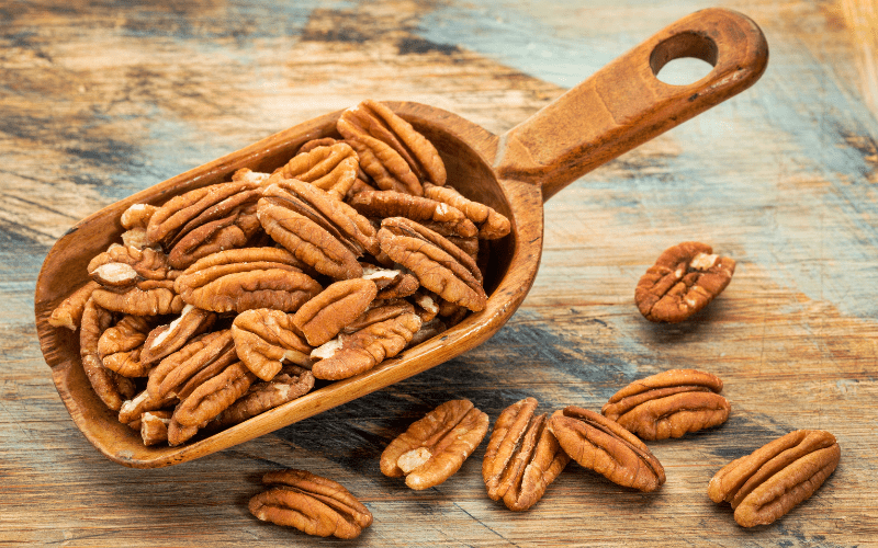 A close-up view of unshelled pecans arranged on a wooden scooper, set on a textured rustic wood table. The warm, earthy tones of the pecans and the natural textures of the wooden surface create a visually appealing composition, evoking a sense of wholesome, natural indulgence.
